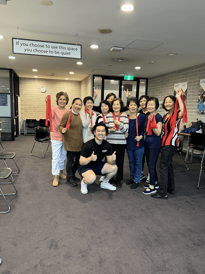 Clinician crouching with a thumbs up in front of a group of older women holding resistance bands in a community room