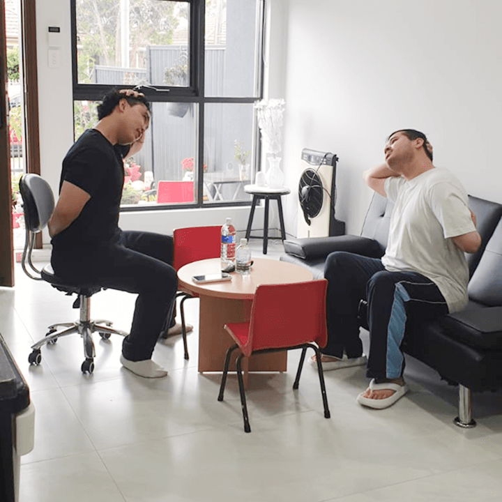 Clinician and participant seated facing each other, both doing a neck stretch with one hand on the head