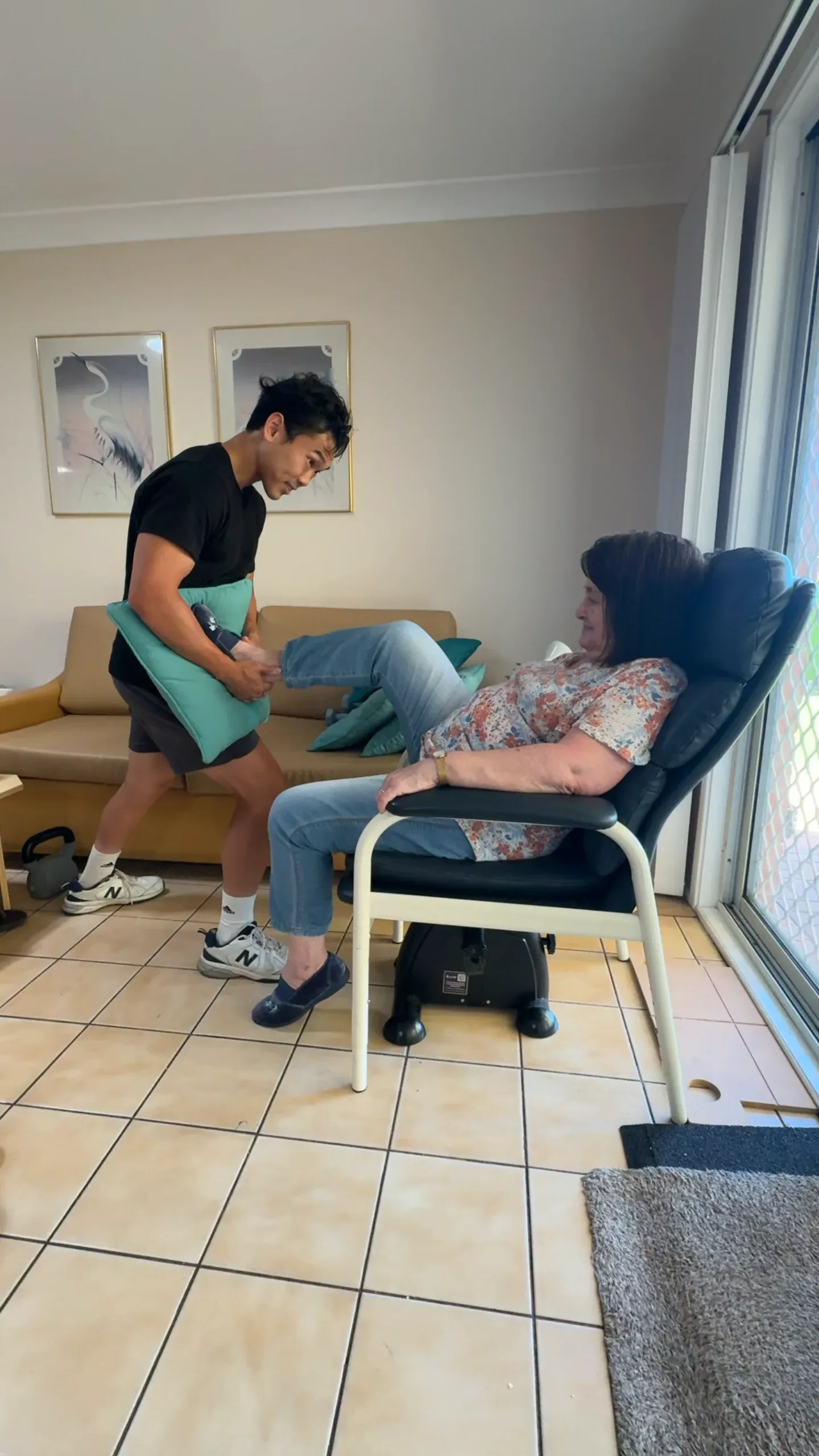 Clinician lifting and guiding an older woman's leg as she sits in a recliner in her living room