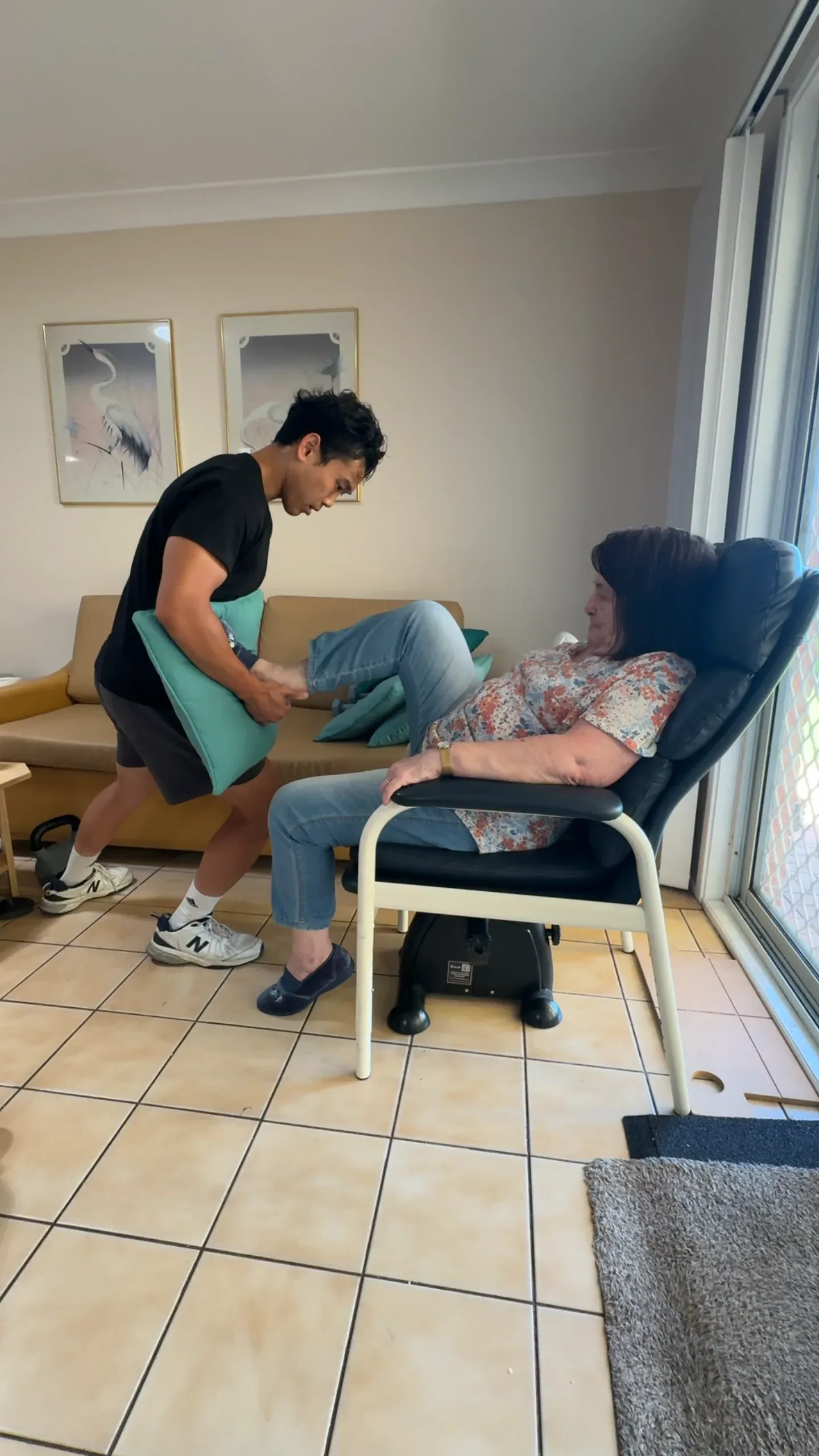 Clinician working with an older woman seated in a recliner in her living room, a cushion under her leg