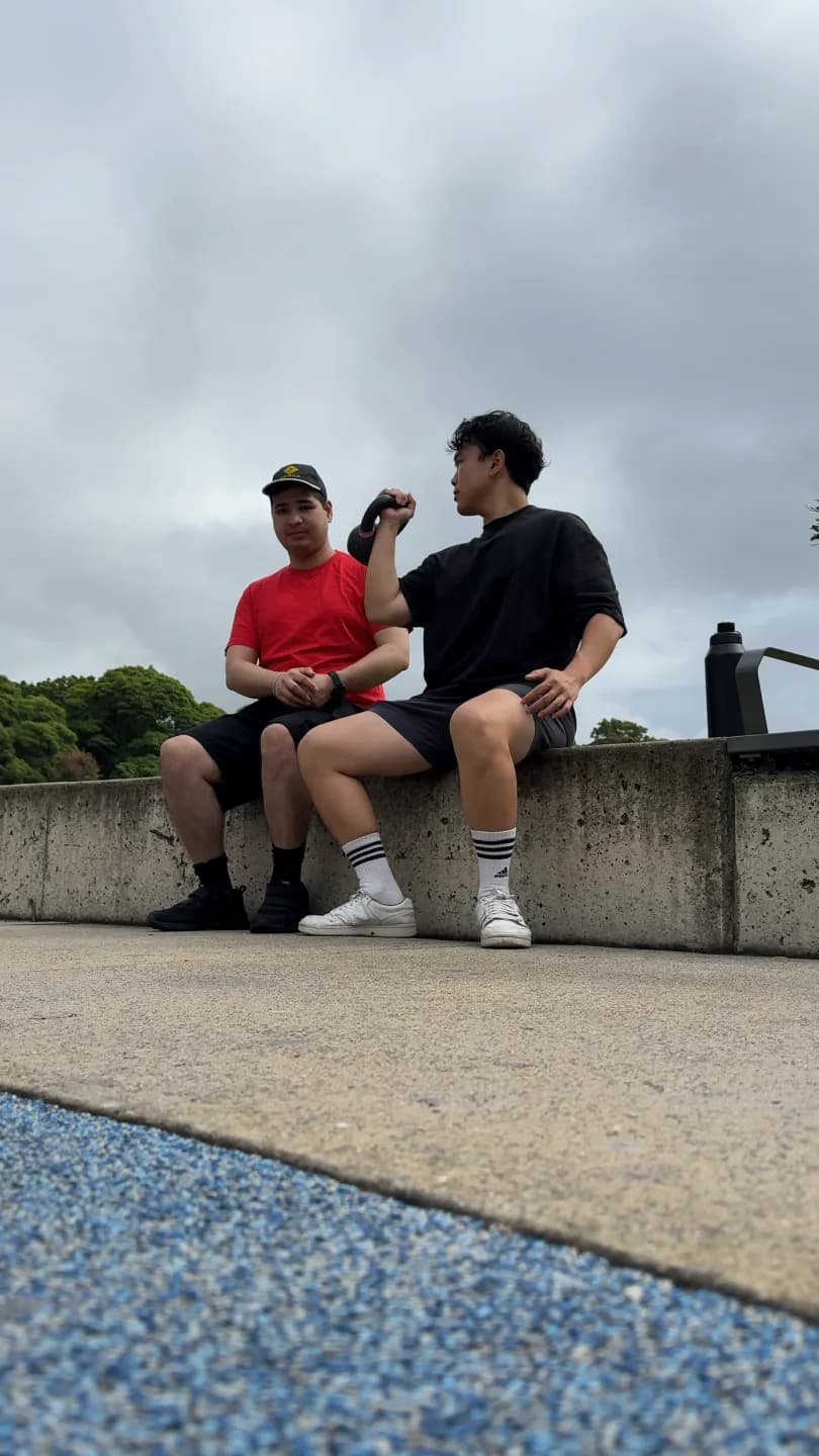 Clinician demonstrating an arm curl with a kettlebell while sitting beside an adult on a low wall outdoors
