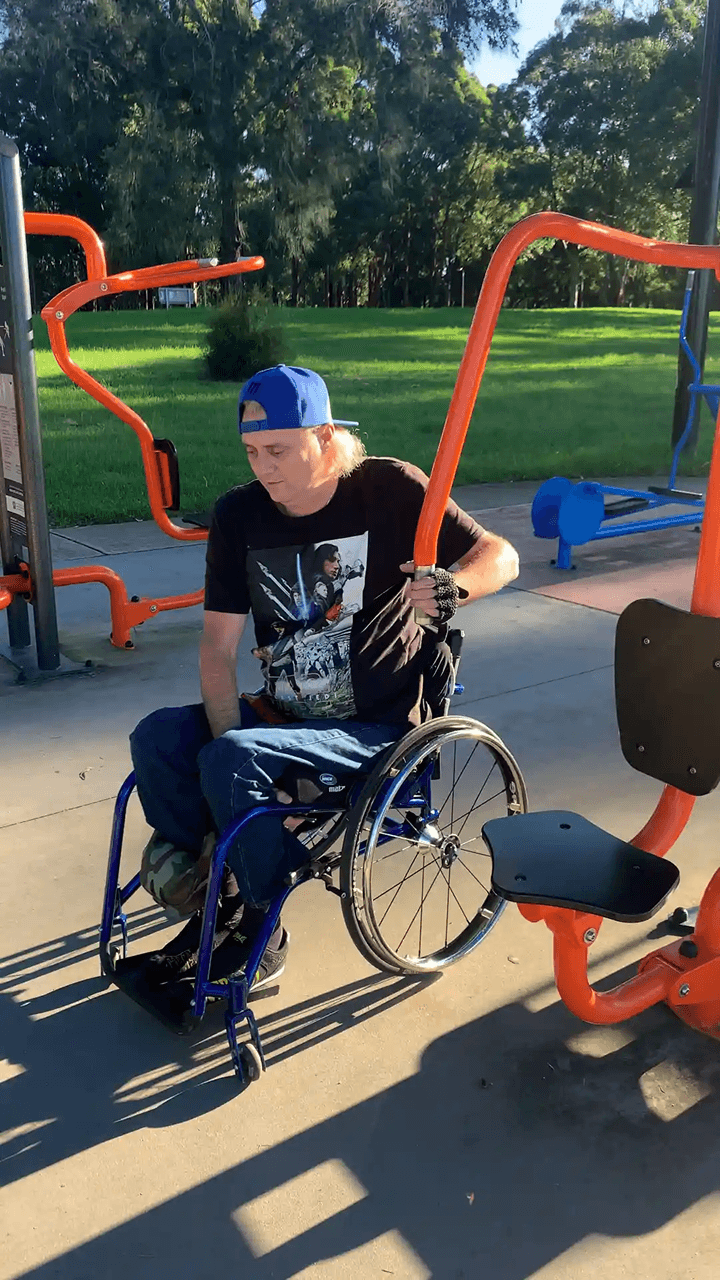 Man in a wheelchair holding the handle of orange outdoor exercise equipment at a park